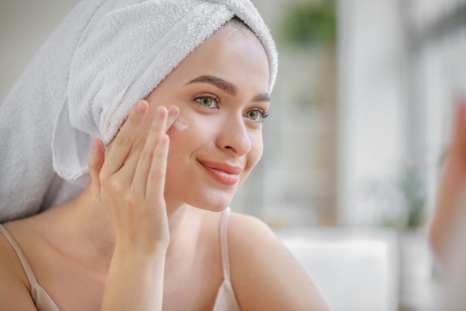 A woman in a towel wrap smiling as she smooths cream onto her cheek